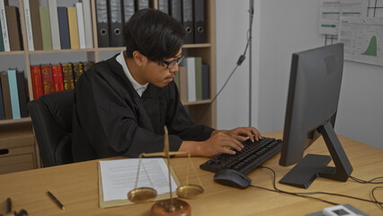 Young man in judicial robe working at his desk in an office with legal books and scales in a professional courtroom setting.