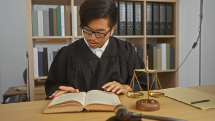 Young, chinese man in a judge's robe reading a book in an office with scales of justice and a gavel on the desk.