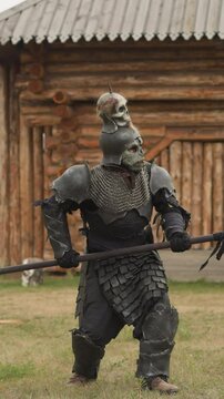 Creepy knight with skull on helmet runs holding spear menacingly rain. Evil warrior rushes to battle moving heavily in medieval settlement with wooden buildings