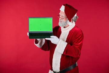 Santa Claus showcasing laptop with green screen ideal for adding custom content. Elderly man standing against red background wearing Santa costume holding laptop