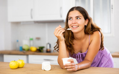 Dreamy girl in a purple nightgown eating delicious yogurt at home kitchen