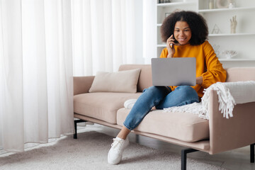 Always In Touch. Happy young african american woman talking on mobile phone, sitting on the couch, using laptop. Copy space, blurred background. Communication Concept. High quality photo