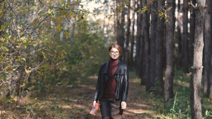 Fototapeta premium Smiling city woman relaxing in the forest. Thoughtful woman in glasses and office clothes walking in the autumn park, breathing fresh air, enjoying nature.