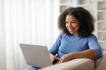 Cheerful young black woman using laptop while sitting on sofa at home, smiling african american female freelancer browsing or working online on computer, typing on keyboard, copy space. High quality