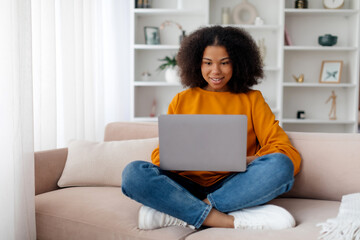 Fototapeta premium Busy cheerful pretty millennial African American female typing on computer in minimalist living room interior. Study, freelance, work and business remotely at home, gadget and new normal. High quality