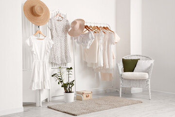 Rack with stylish female clothes, hats, wicker armchair and houseplant near light wall in room