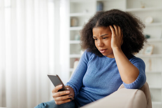 Concerned young black woman looking at smartphone screen and touching head, upset african american female sitting on couch with troubled expression, suffering stress or got bad news on phone. High