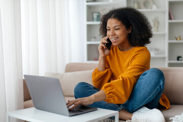 Online Work Concept. Young Black Woman Working With Laptop And Talking On Cellphone At Home, African American Woman Using Computer For Freelancing, Sitting On Couch in Living Room, Copy Space. High