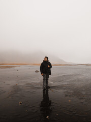 Photographer walking on wet sand near a lake in iceland on a cloudy day with mountains in the background