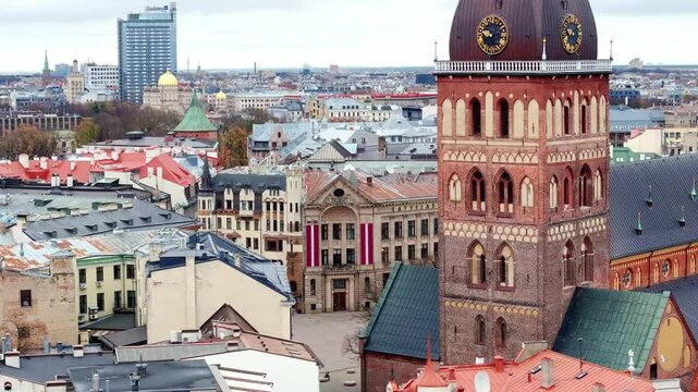 The image shows an aerial view of Riga, Latvia, highlighting the Riga Cathedral and its clock tower. Modern and historical buildings, including the Radisson Blu hotel, are visible.