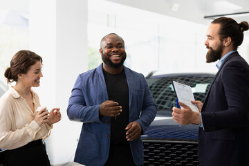 Funny car salesman in dealership making clients laugh to help solidify deal, earning commission...