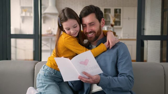 Smiling carefree Caucasian dad man parent reading card present from little daughter kid. Loving family male father guy hugging girl child touch at home Father's Day celebration gift parenting holiday