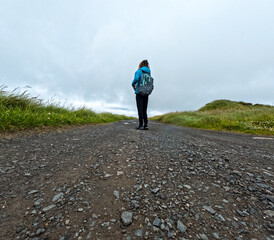 Rear view of a single female backpacker hiking on a gravel road. She is dressed casually and takes in the beautiful views of nature. Nólsoy, Faroe Islands.