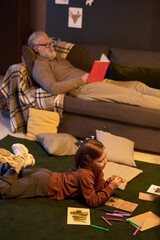 Fototapeta premium Senior man relaxing on couch reading book with child lying on floor drawing. Cozy home environment with pillows scattered around room creating warm atmosphere