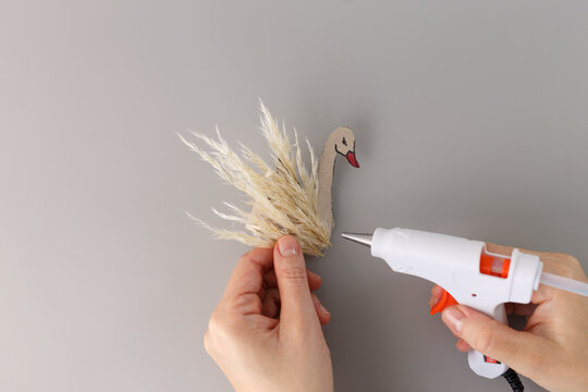 Hands crafting swan figurine from cardboard and pampas grass using hot glue gun on a grey background. DIY decoration project concept. Design for tutorial, craft blog, creative hobby content,