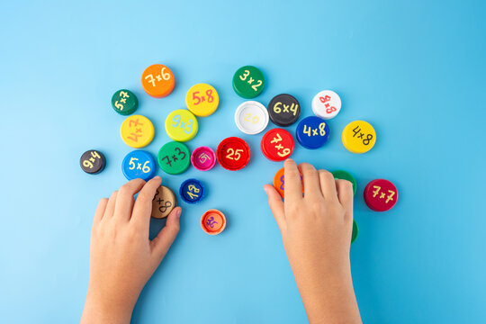 multiplication table learning technique for kids, studding through the game, hands arranging colorful bottle caps with multiplication problems on a blue background. Flat lay educational concept ,
