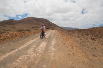 a middle aged woman in a cowboy hat riding a bicycle through a desertic landscape, horizontal, La Graciosa, Lanzarote