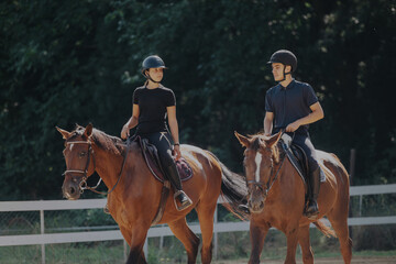 Young couple riding horses in an outdoor setting. They are wearing helmets and casual attire,...