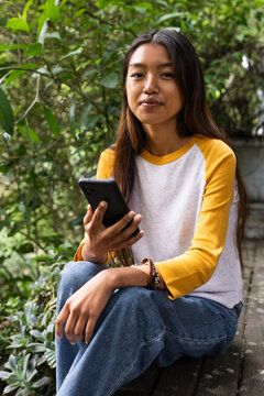 Sitting outdoors, asian female teenager holding smartphone and appreciating nature and technology
