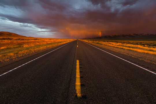Endless summer desert highway leading to the horizon at sunset with dramatic storm clouds and golden hour light in Arizona and Utah