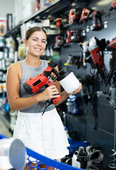 Fototapeta premium Girl buyer inspects modern power tool in construction hypermarket and selects picks up spray gun. Young woman near showcase with construction equipment