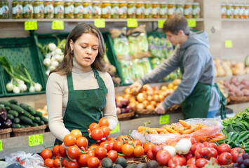 Young woman seller in apron lays out tomatoes on counter in vegetable shop