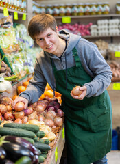 Young salesman in apron lays out onions on counter in vegetable shop
