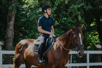 A young equestrian riding a brown horse in a sunny outdoor arena, surrounded by lush green trees....