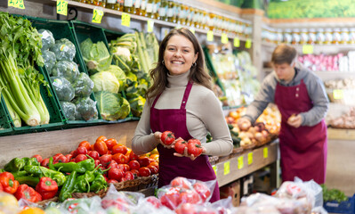 Young woman seller in uniform lays out tomatoes on counter in grocery store
