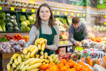 Positive young female seller holding bananas standing by counter in fruit and vegetable market