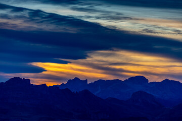 Sunset in the mountains. Red and orange burning sky horizon behind the alpine mountain range. Rocky mountain peaks of the Dolomites at sunset and dawn at sunrise light sunrays going through the clouds