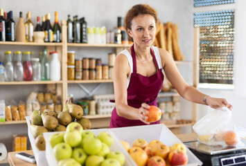 Adult saleswoman weighing apples in bag on scales in grocery store