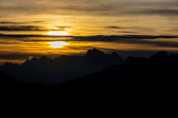 Sunset in the mountains. Red and orange burning sky horizon behind the alpine mountain range. Rocky mountain peaks of the Dolomites at sunset and dawn at sunrise light sunrays going through the clouds