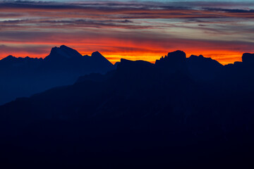 Sunset in the mountains. Red and orange burning sky horizon behind the alpine mountain range. Rocky mountain peaks of the Dolomites at sunset and dawn at sunrise light sunrays going through the clouds