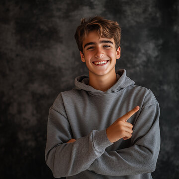 A joyful 16-year-old boy with a bright smile, standing proudly against a dark textured background, wearing a casual gray hoodie, and pointing confidently to the side.