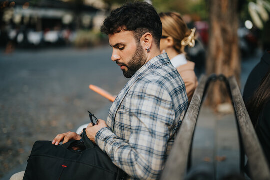 A young businessman sits on a park bench outdoors, holding his smart phone. He appears focused and professional, reflecting a modern lifestyle. The scene exudes a casual yet business-oriented - Powered by Adobe