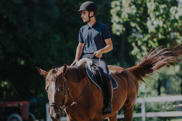 A young man, fully equipped with riding gear, enjoys a serene horseback ride in a lush green...