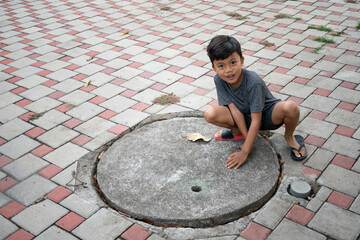 A curious young Asian boy looking at the camera while crouching down on a patterned brick sidewalk,...