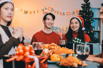 Group of young Asian man and women as friends having fun at a New Year's celebration, holding gift boxes standing by Christmas tree decoration, midnight countdown Party at home with holiday season.