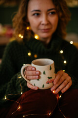 Mid-aged woman sitting indoors, holding a Christmas-themed mug and surrounded by warm holiday lights, creating a cozy and serene Christmas atmosphere