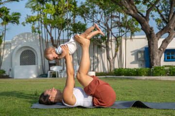 In a tropical paradise, a woman practices yoga on fresh grass, playfully lifting her baby girl...