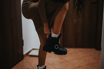 A young woman is seen adjusting her stylish black boot, capturing a moment of everyday fashion indoors. The scene conveys a sense of modern style and casual elegance.