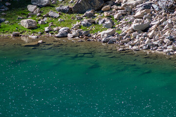 Clear turquoise water gently laps against the rocky shore of a serene lake surrounded by greenery on a sunny day