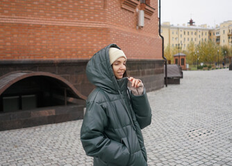 A joyful woman wearing a green puffer jacket and a beanie stands in a park, with colorful autumn...
