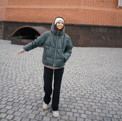 A joyful woman wearing a green puffer jacket and a beanie stands in a park, with colorful autumn...