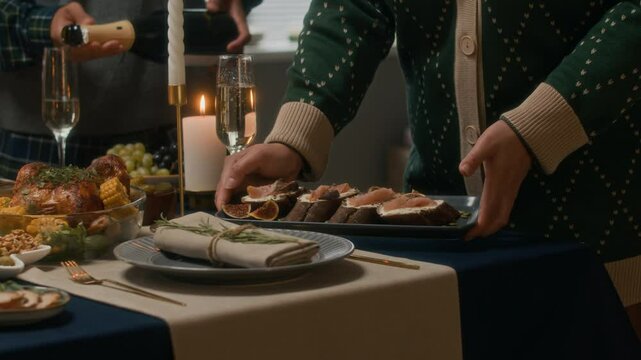 Cropped slowmo shot of two unrecognizable hospitable people serving appetizers on elegant tableware and pouring champagne in glasses while making final table setting preparations for Christmas