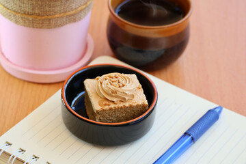 A small light brown cake on a notebook with a pen on it. Next to it is a cup of hot brown coffee at a wooden table.