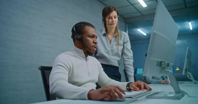 Modern call center office: Two multiethnic technical support specialists wearing headsets talk, discuss work monitoring data on computer. Team of hotline operators working in online customer support.