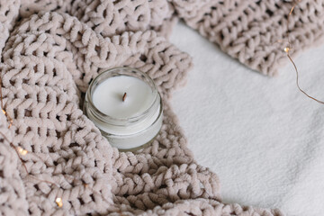 A candle in a glass jar lies among a knitted plush soft warm plaid. Warm comfort