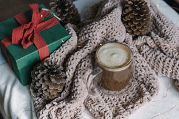 Winter still life. A glass cup of coffee with milk, big cones, lights, a green gift box with a red bow. Modern christmas rustic eco friendly decor, scandinavian winter hygge. Atmospheric christmas